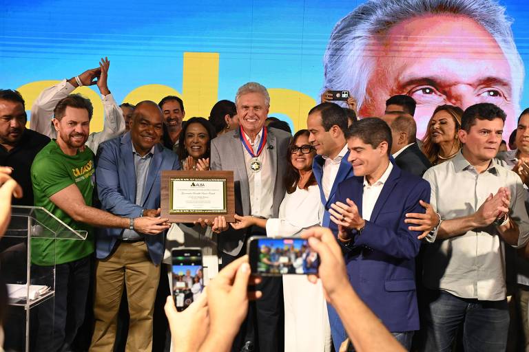 A imagem mostra um grupo de pessoas em uma cerimônia de premiação. No centro, um homem de cabelo grisalho, usando uma medalha, segura um quadro com uma placa. Ao redor dele, várias pessoas aplaudem e sorriem, algumas segurando celulares para registrar o momento. O fundo é colorido, com um grande painel que parece ter a imagem de um rosto e a palavra 039;Agradecimento039;.