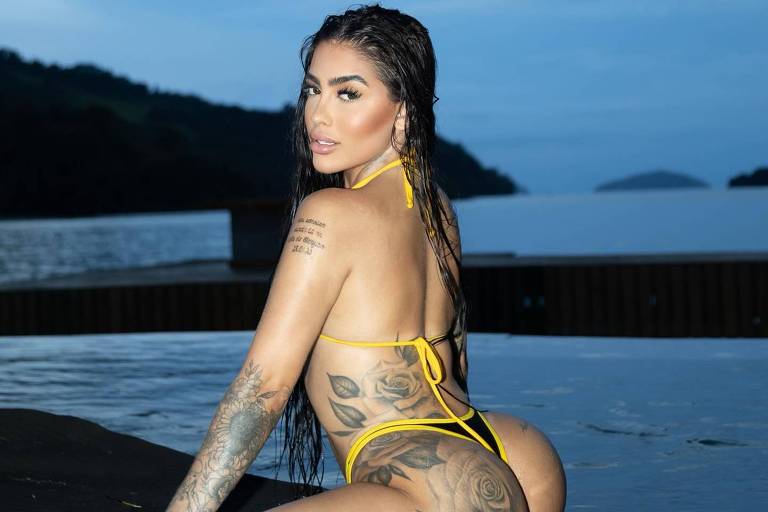 Uma mulher com cabelo molhado e tatuagens está sentada em uma prancha de surf, em uma água calma. Ela usa um biquíni amarelo e está voltada para a câmera, com um fundo de céu nublado e montanhas ao longe.