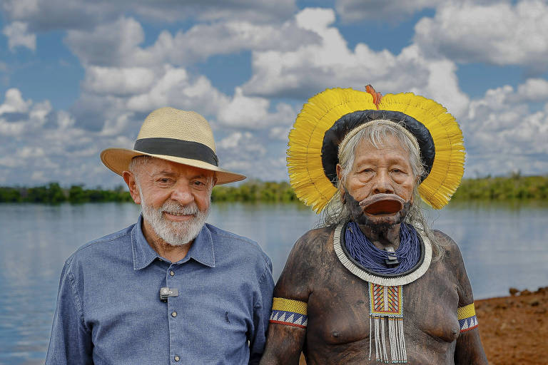 A imagem mostra um homem idoso com cabelo grisalho e barba, usando uma camisa azul e um chapéu de palha, ao lado de uma mulher indígena com pintura corporal, adornos de penas amarelas e um colar. Ao fundo, há um rio e um céu com nuvens brancas.