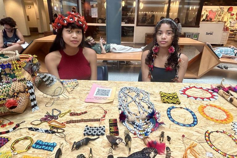 Duas jovens sentadas em uma mesa com diversos itens de artesanato expostos, incluindo colares, pulseiras e outros acessórios. Uma das jovens usa uma coroa de flores, enquanto a outra tem brincos grandes. Ao fundo, há pessoas sentadas e um ambiente de feira.