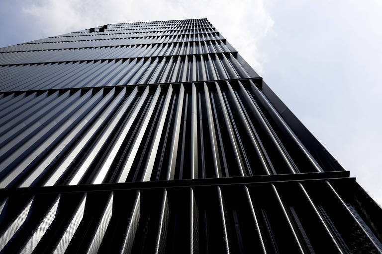 A imagem mostra a fachada de um edifício alto, com uma superfície preta e texturizada, composta por painéis verticais e linhas retas. A perspectiva é de baixo para cima, destacando a altura do edifício sob um céu parcialmente nublado.