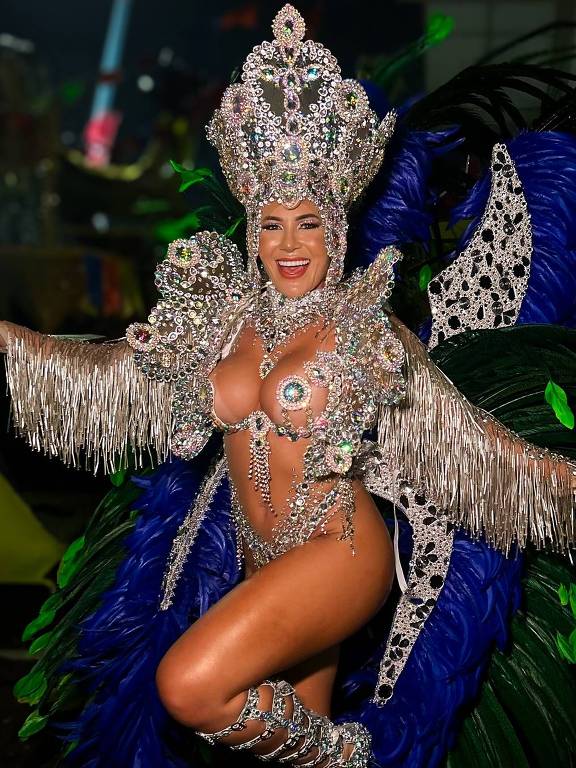 Uma mulher está posando em um evento de carnaval, vestindo uma fantasia elaborada com muitos detalhes. A fantasia é adornada com pedras brilhantes e plumas azuis, além de ter franjas prateadas. Ela está sorrindo e levantando uma perna, enquanto usa sandálias com tiras brilhantes. O fundo é desfocado, mas sugere um ambiente festivo.