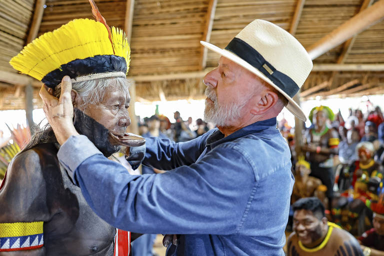 A imagem mostra um homem de cabelos grisalhos e barba, usando um chapéu claro, interagindo com um indígena que está vestido com trajes tradicionais e uma máscara. O ambiente é uma cerimônia indígena, com várias pessoas ao fundo, algumas vestindo roupas coloridas e adornos típicos. O local parece ser uma estrutura de madeira com um teto de palha.