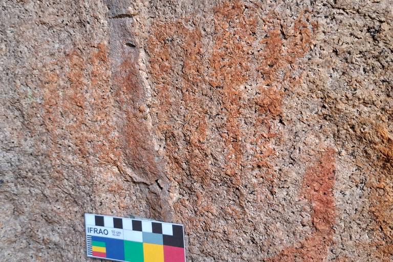 A imagem mostra uma superfície de rocha com várias marcas de pinturas rupestres em tons de laranja e vermelho. Na parte inferior, há uma régua de escala colorida para referência de tamanho.