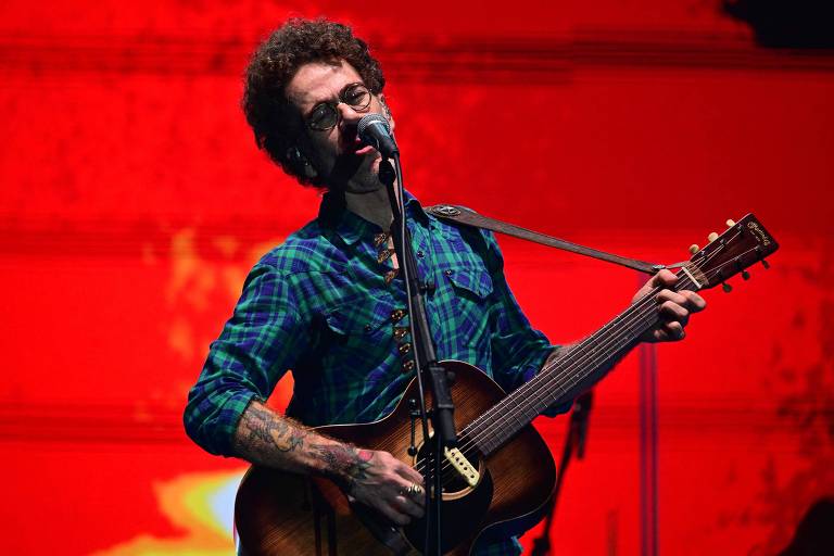 Um músico está se apresentando ao vivo, cantando em um microfone enquanto toca violão. Ele usa uma camisa xadrez azul e verde e possui cabelos cacheados e barba. O fundo da imagem é de uma cor vermelha vibrante, criando um contraste com o artista.