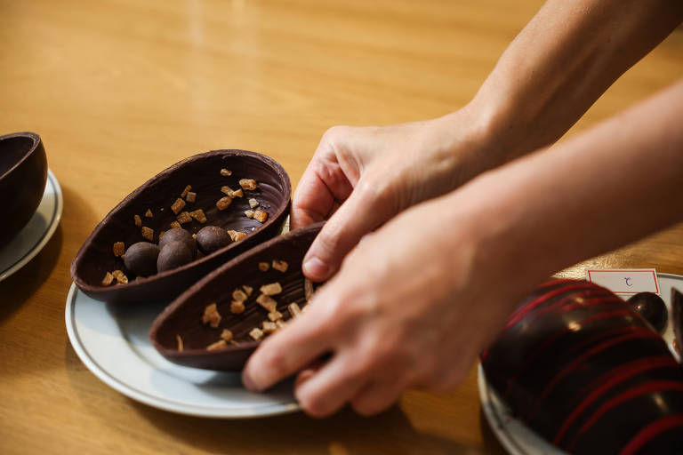 A imagem mostra uma mão segurando uma casca de chocolate em formato oval, que contém bolinhas de chocolate e pedaços de caramelo. Ao lado, há outra casca de chocolate decorada com listras vermelhas. O fundo é uma mesa de madeira clara.