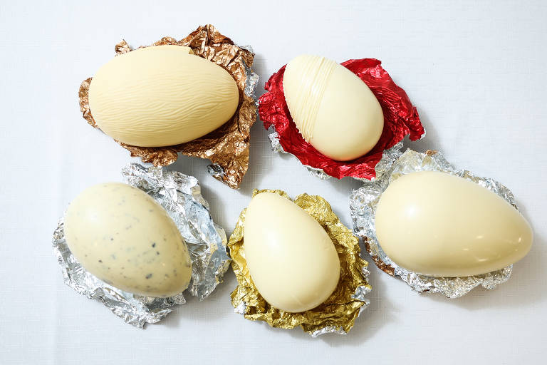 A imagem mostra cinco ovos de chocolate dispostos sobre papéis coloridos. Os ovos são de diferentes cores e texturas, incluindo um ovo marrom claro com uma textura de madeira, um ovo branco liso, um ovo com manchas escuras, e outros em tons de creme e dourado. Os papéis que envolvem os ovos são de cores variadas, como vermelho, dourado e prateado.
