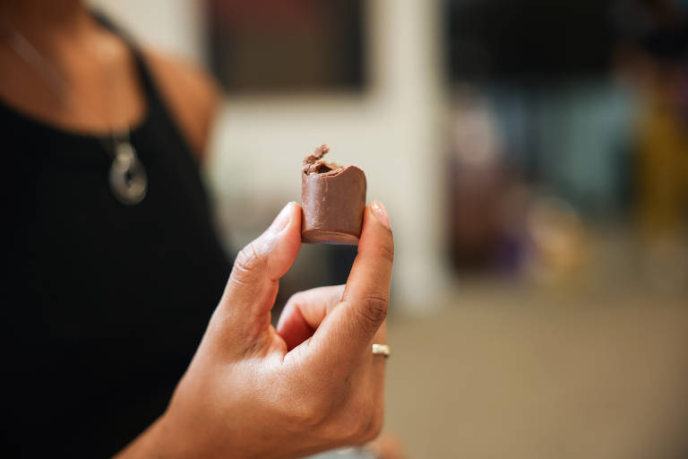 Uma pessoa está segurando um pedaço de chocolate com uma mordida. O chocolate é de formato pequeno e parece ser cremoso. A mão que segura o chocolate é visível, e a pessoa está usando uma blusa preta. O fundo é desfocado, com algumas cores indistintas.