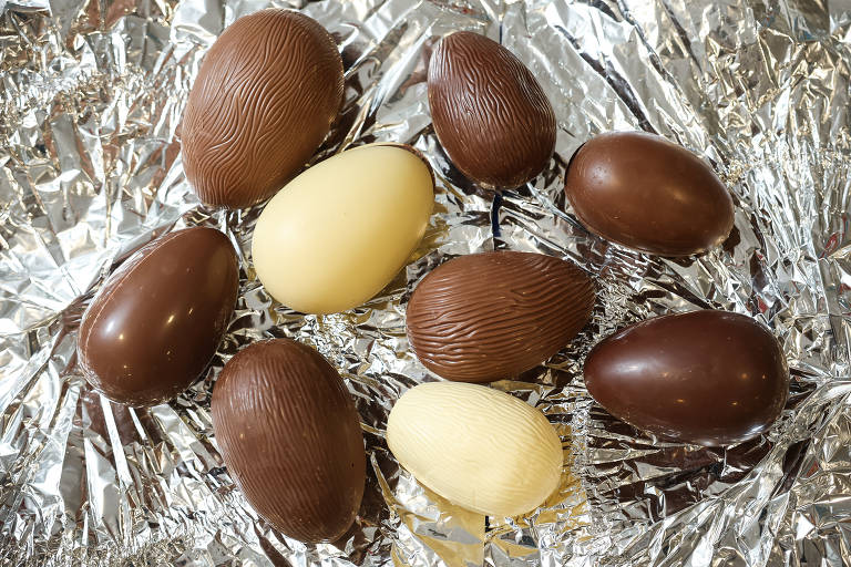 A imagem mostra uma variedade de ovos de chocolate dispostos sobre um fundo de papel alumínio. Existem cinco ovos de chocolate ao leite, três ovos de chocolate branco e dois ovos de chocolate escuro, todos com texturas diferentes na superfície.