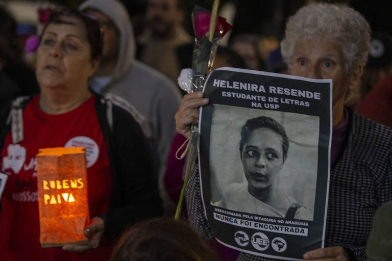 A imagem mostra um grupo de pessoas em uma manifestação. À frente, uma mulher segura um cartaz com a foto de Helenira Resende, que é identificada como estudante de Letras na USP, e a frase 'NUNCA FOI ENCONTRADA'. Outra mulher, à esquerda, segura uma lanterna com o nome 'RUBENS PAIVA' e tem uma rosa na mão. O ambiente parece ser noturno, com outras pessoas ao fundo.