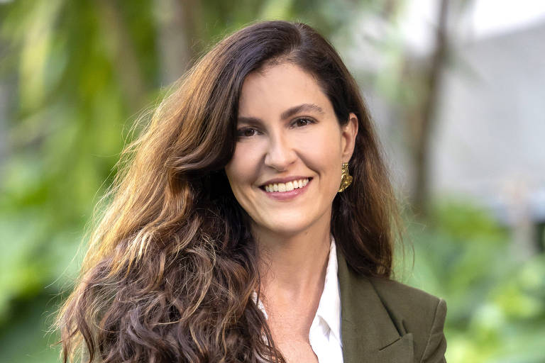 Uma mulher com cabelo longo e ondulado, sorrindo para a câmera. Ela está vestindo um blazer verde e uma blusa branca. Ao fundo, há uma vegetação densa, com folhas verdes e um ambiente natural.