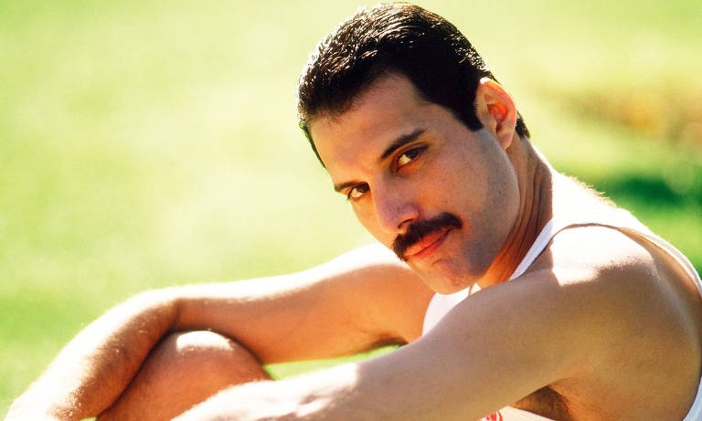 A imagem mostra um homem com cabelo escuro e um bigode, sentado em um ambiente ao ar livre. Ele está usando uma camiseta sem mangas e olhando diretamente para a câmera com uma expressão suave. O fundo é desfocado, com grama verde.