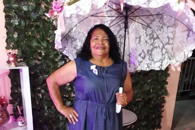 A imagem mostra uma mulher sorridente em pé, segurando um guarda-chuva aberto. Ela está vestindo um vestido azul com bordados coloridos na parte inferior. O fundo é decorado com plantas verdes e uma estante branca com objetos decorativos. O ambiente tem iluminação suave e um tapete vermelho.