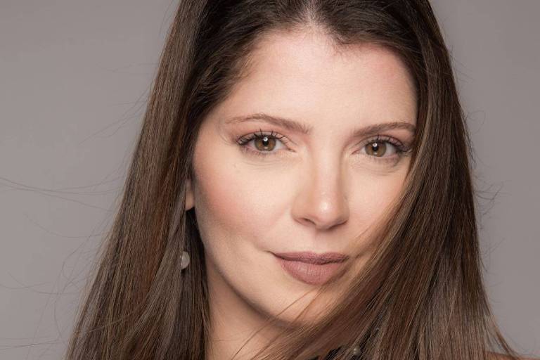 A imagem mostra uma mulher com cabelo longo e liso, que cai sobre os ombros. Ela está usando uma blusa preta com detalhes em pérolas. O fundo é neutro e a iluminação é suave, destacando seu rosto e expressão confiante.