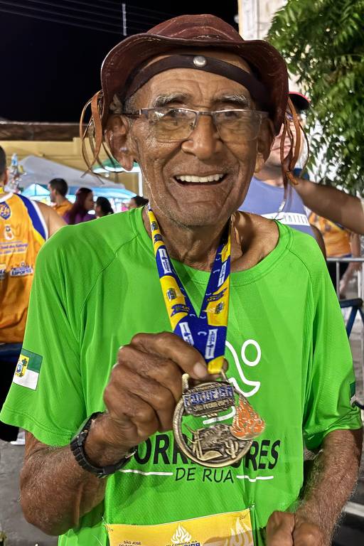 Um homem idoso, sorridente, usando uma camiseta verde com a inscrição 'CORREDORES DE RUA', está segurando uma medalha em sua mão. Ele usa um chapéu marrom e óculos. Ao fundo, há outras pessoas e barracas, sugerindo um evento ao ar livre.