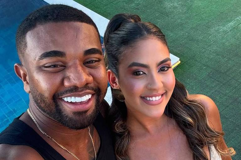 A imagem mostra um casal tirando uma selfie. O homem à esquerda está usando uma camiseta preta e sorrindo, enquanto a mulher à direita está vestindo um vestido branco com detalhes em crochê e também sorri. Ao fundo, há uma área verde e uma piscina.