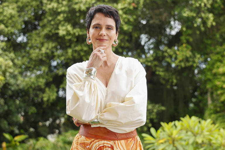 Uma mulher está em pé em um ambiente ao ar livre, com árvores e plantas ao fundo. Ela usa uma blusa branca de mangas largas e uma saia laranja com padrões. A mulher tem cabelo curto e escuro, e está com uma expressão pensativa, apoiando o queixo com a mão. Ela usa brincos grandes e uma pulseira prateada.