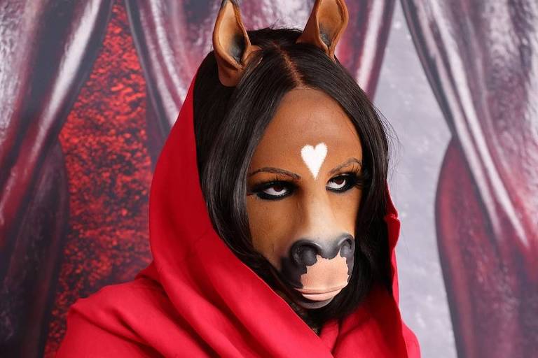 A imagem mostra uma pessoa usando uma máscara que imita a cabeça de um cavalo, com um coração branco na testa. A pessoa está vestida com um manto vermelho que cobre parte do corpo. O fundo da imagem é composto por cortinas vermelhas, criando um contraste com a vestimenta e a máscara.