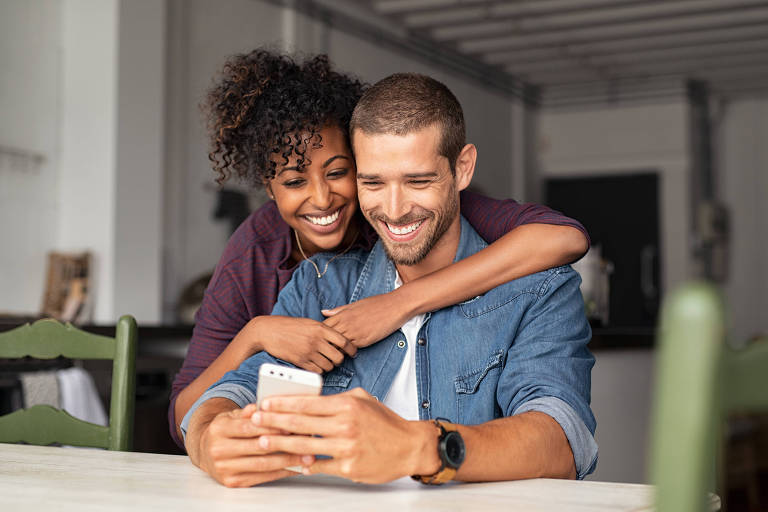 Um casal está sentado à mesa, sorrindo enquanto olha para um smartphone. A mulher, com cabelo cacheado e vestido escuro, está abraçando o homem, que tem cabelo curto e usa uma camisa jeans. Ambos parecem felizes e envolvidos na atividade que estão fazendo juntos.