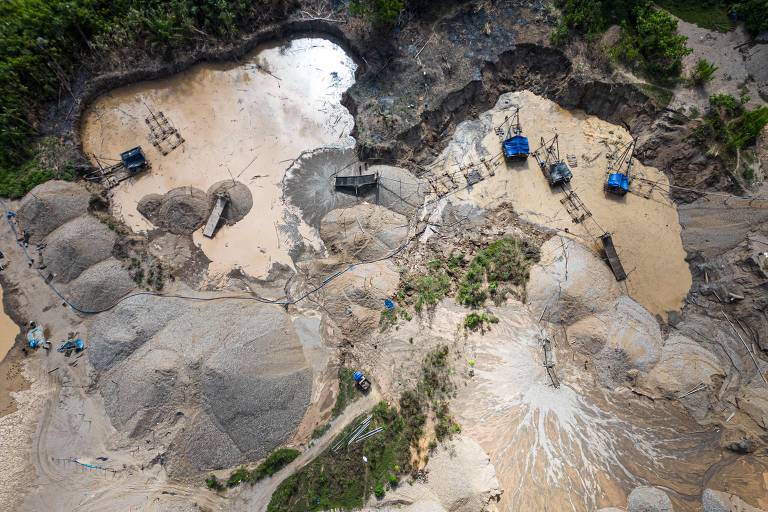 A imagem mostra uma área de mineração em um ambiente fluvial, com várias pilhas de areia e cascalho. Há também máquinas de mineração, como caminhões, visíveis na cena. O solo está molhado e há poças de água, indicando a presença de um corpo d'água nas proximidades. A vegetação ao redor é densa, sugerindo que a área é uma floresta tropical.