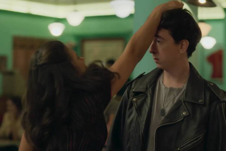 A imagem mostra uma cena em um ambiente social, possivelmente um restaurante ou bar. Uma mulher com cabelo longo e solto está levantando a mão em direção à cabeça de um homem, que está usando uma jaqueta de couro preta. O homem parece surpreso ou confuso. O fundo é decorado com paredes verdes e luzes suaves, e há outras pessoas visíveis ao fundo, sugerindo um ambiente animado.