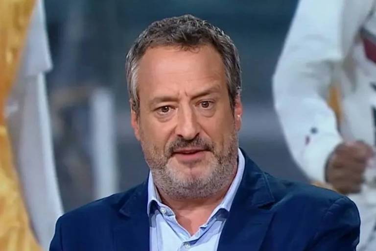 Um homem com barba e cabelo grisalho está falando em um programa de televisão. Ele usa um paletó azul e uma camisa clara. Ao fundo, há uma figura desfocada vestindo um traje colorido. O ambiente parece ser um estúdio de TV.