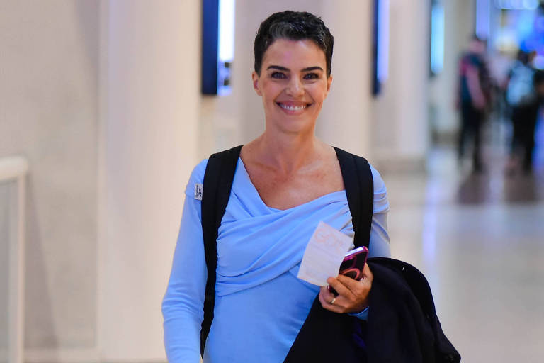 Uma mulher com cabelo curto e escuro, vestindo uma blusa azul clara, está sorrindo enquanto caminha em um ambiente interno. Ela carrega uma bolsa preta e um celular na mão, e parece estar em um espaço público, possivelmente um aeroporto ou shopping, com pessoas ao fundo.