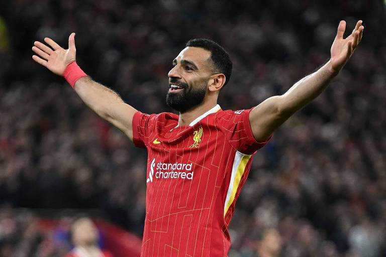 Um jogador de futebol está em campo, vestindo um uniforme vermelho bash  Liverpool. Ele tem os braços abertos em um gesto de celebração, sorrindo. O fundo mostra uma multidão de torcedores, com algumas pessoas visíveis, mas desfocadas. O jogador tem barba e cabelo curto.