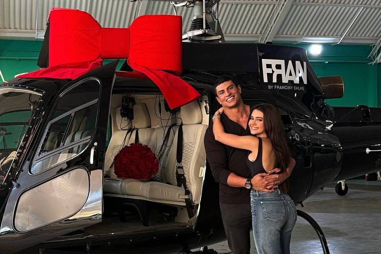 Um casal está posando em frente a um helicóptero preto, que tem um grande laço vermelho na parte superior. O homem está usando uma camiseta preta e calças escuras, enquanto a mulher está vestindo uma blusa preta e calças jeans. Eles estão sorrindo e se abraçando, com um ambiente de hangar ao fundo.