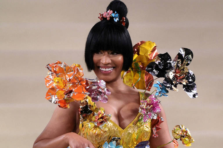 Uma mulher está desfilando em uma passarela, vestindo um vestido curto e brilhante, predominantemente amarelo, decorado com flores coloridas feitas de papel ou material similar. Ela tem cabelo preto liso e usa um coque alto, além de acessórios como anéis. A expressão dela é sorridente e confiante, enquanto ela segura uma pequena bolsa. O fundo é neutro, destacando a modelo e o vestido.