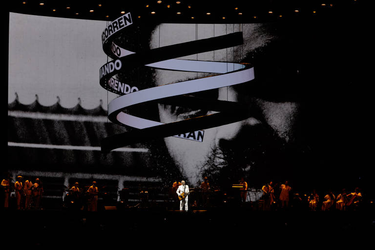 A imagem mostra um palco de apresentação musical com um grande telão ao fundo. No telão, há uma projeção em preto e branco de um rosto, possivelmente de um homem, com texto em letras grandes que diz "CORREN DO" e "ANDANDO". No palco, há vários músicos e um artista em destaque, vestido de branco, que parece estar se apresentando. O ambiente é iluminado com luzes focadas no artista e na orquestra.