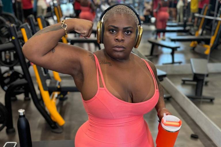 Uma mulher com cabelo raspado está em uma academia, usando um macacão rosa e fones de ouvido. Ela está fazendo um gesto de força com o braço, enquanto segura um copo laranja na outra mão. Ao fundo, há várias pessoas e equipamentos de musculação.
