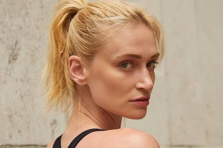 A imagem mostra uma mulher com cabelo loiro preso em um coque alto, usando um top preto. Ela está de perfil, olhando para trás, e exibe várias tatuagens nos braços. O fundo é de uma parede clara e texturizada.