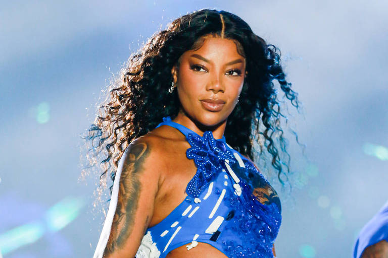 Uma artista feminina está se apresentando em um palco. Ela tem cabelo longo e cacheado, e usa um traje azul que é composto por um top e uma saia. O fundo é iluminado com luzes coloridas, criando uma atmosfera vibrante de show ao vivo.