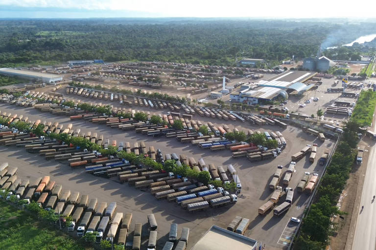 A imagem mostra uma grande área de estacionamento para caminhões, com muitos veículos dispostos em fileiras organizadas. O local é cercado por vegetação e há um edifício visível ao fundo, possivelmente um terminal ou centro de serviços. À direita, uma estrada pode ser vista, com caminhões também estacionados ao longo dela. O céu está claro e há uma vista de um rio na parte superior da imagem.