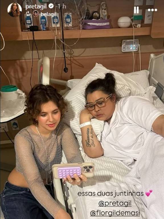 Duas mulheres estão sentadas em uma cama de hospital. Uma delas está deitada, usando uma blusa branca e óculos, enquanto a outra está ao lado, usando uma blusa transparente e jeans. Ambas estão olhando para um celular que está na frente delas. O ambiente é de hospital, com equipamentos médicos visíveis ao fundo. Há uma legenda que diz 'essas duas juntinhas' e menciona perfis de redes sociais.