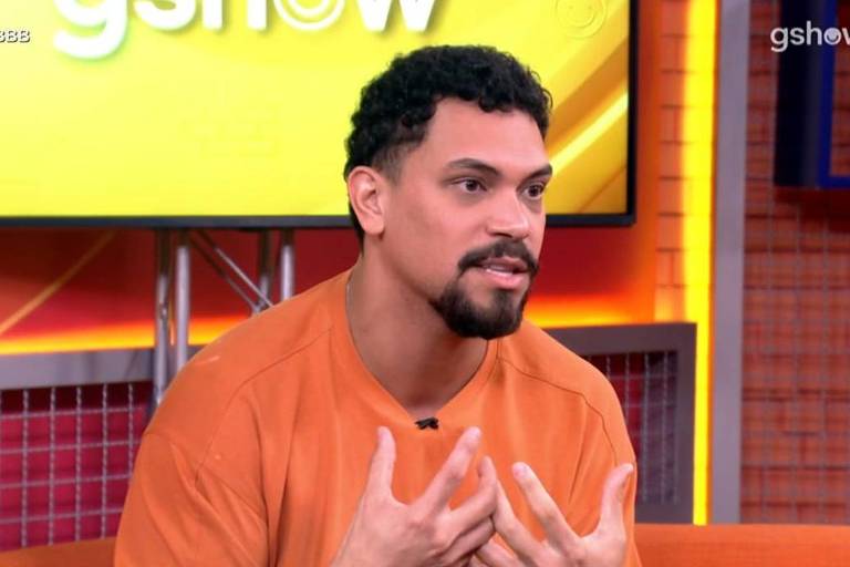 Um homem com cabelo cacheado e barba está sentado em um estúdio, vestindo uma camiseta laranja. Ele parece estar falando e gesticulando com as mãos, enquanto uma tela amarela ao fundo exibe o logo do programa. O ambiente é colorido e moderno, com elementos decorativos visíveis.