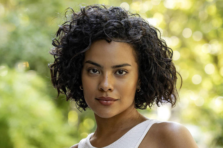 A imagem mostra uma mulher com cabelo cacheado e escuro, vestindo uma blusa branca sem mangas e calças jeans azuis. Ela está em pé em um ambiente ao ar livre, com plantas ao fundo, e olha diretamente para a câmera com uma expressão neutra.