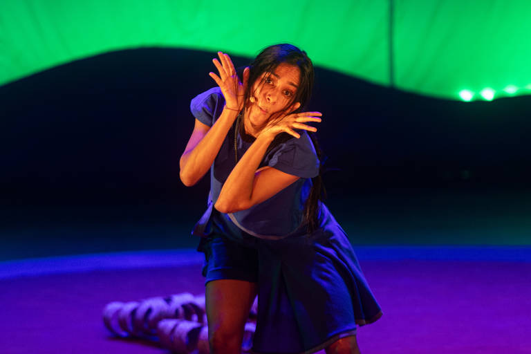 Uma artista de performance está em cena, com uma expressão facial expressiva e gestos dramáticos. Ela usa uma roupa azul e está em um palco iluminado com um fundo verde. O chão do palco é de cor roxa, e há uma figura enrolada em tecido ao fundo.