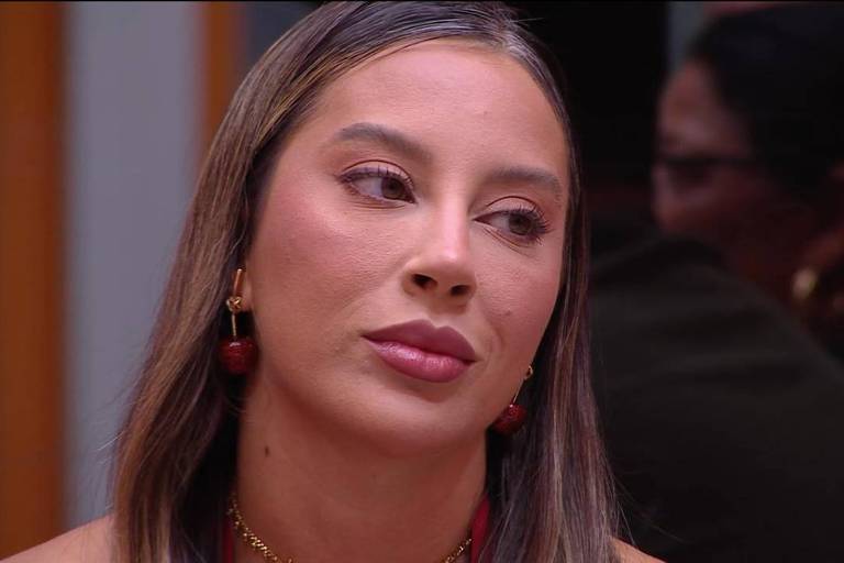 A imagem mostra uma mulher com cabelo longo e liso, usando brincos vermelhos e um colar dourado. Ela tem uma expressão pensativa e está olhando para o lado, em um ambiente que parece ser uma reunião ou evento social. Ao fundo, é possível ver outras pessoas, mas não estão claramente visíveis.