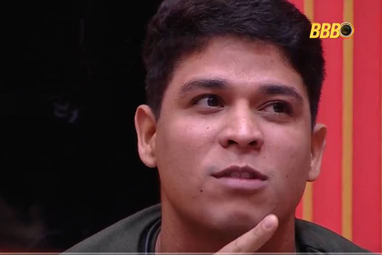 Um homem com cabelo curto e pele clara está sentado, olhando para o lado com uma expressão pensativa. Ele está usando uma camiseta escura e tem uma mão apoiada no queixo. Ao fundo, há uma parede com listras vermelhas e amarelas, e parte de uma pessoa não identificada é visível à esquerda.