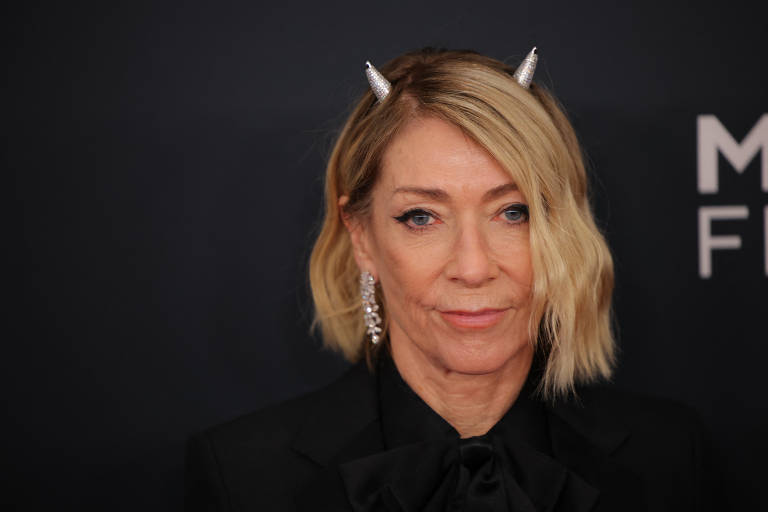A imagem mostra uma mulher com cabelo loiro e ondulado, usando um traje preto. Ela tem chifres prateados na cabeça e brincos grandes. O fundo é escuro, com a palavra 'FUTURO' visível em letras brancas.