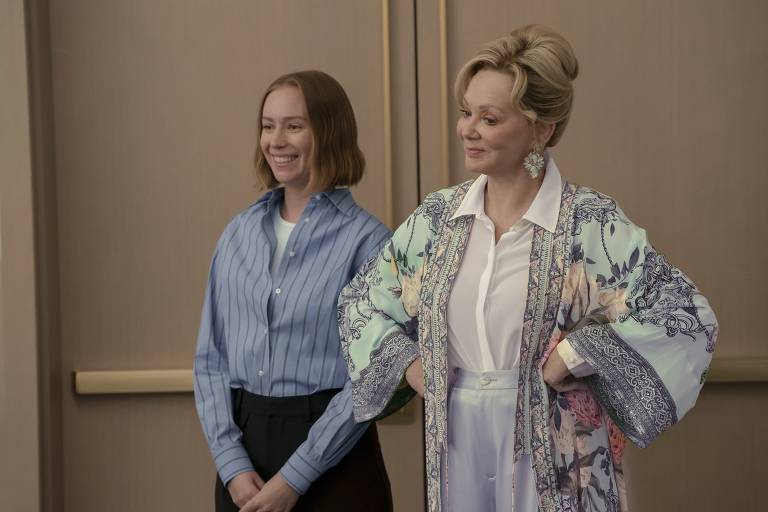 A imagem mostra duas mulheres em um ambiente interno. A mulher à esquerda tem cabelo curto e liso, usa uma camisa azul listrada e está sorrindo. A mulher à direita tem cabelo loiro e usa um robe colorido com estampas florais, além de brincos grandes. Ambas estão em pé, com a mulher à direita com as mãos na cintura e a mulher à esquerda com as mãos juntas na frente do corpo.