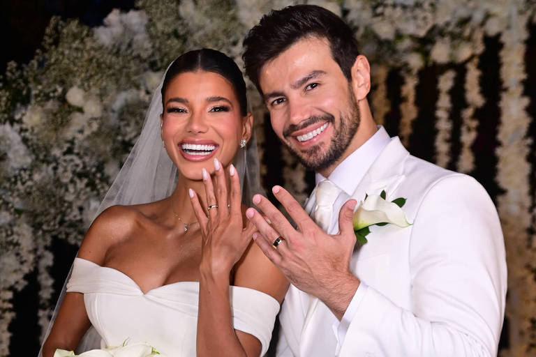 Um casal sorridente posando para a foto após a cerimônia de casamento. A noiva está vestida com um vestido branco elegante, com um decote em coração e segurando um buquê de flores brancas. O noivo está usando um terno branco, com uma flor na lapela. Ambos estão mostrando suas alianças e estão em um ambiente decorado com flores brancas ao fundo.
