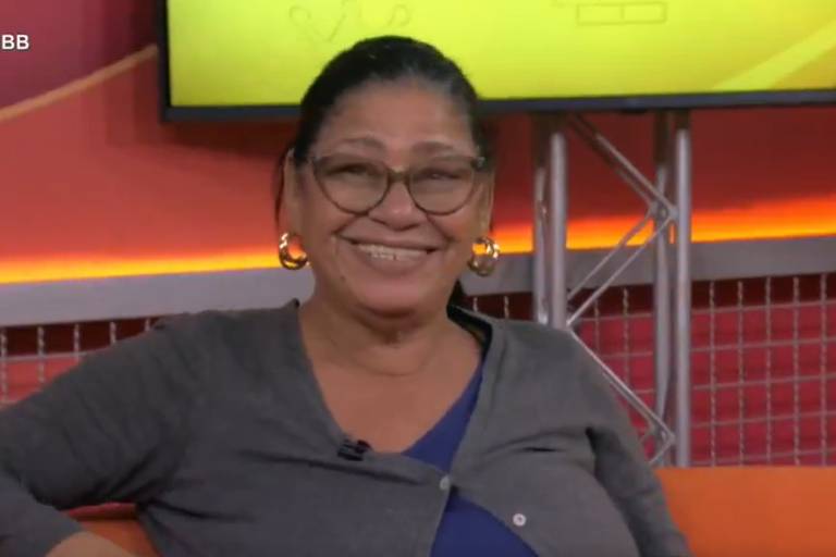 Uma mulher sorridente está sentada em um sofá laranja em um estúdio de televisão. Ela usa óculos e brincos grandes. Ao fundo, há uma tela com um design amarelo e elementos gráficos. A iluminação é suave e o ambiente é acolhedor.