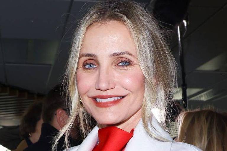 A imagem mostra uma mulher com cabelo loiro e liso, usando um casaco branco e um lenço vermelho. Ela está sorrindo e parece estar em um ambiente interno, com pessoas ao fundo. A iluminação é clara e destaca seu rosto.