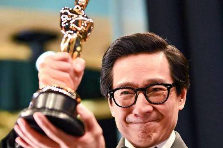 Um homem sorridente segura um troféu do Oscar com uma mão, enquanto a outra mão é visível ao fundo. Ele está usando um terno e gravata, e parece emocionado. O fundo é desfocado, mas sugere um ambiente de premiação.
