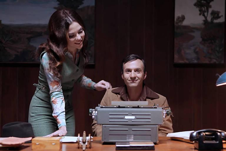 A imagem mostra um homem sentado em uma mesa de escritório, sorrindo, enquanto uma mulher está ao seu lado, inclinando-se levemente em sua direção. O homem está usando uma camisa clara e um casaco marrom, e a mulher está vestida com um vestido verde e uma blusa colorida. Na mesa, há uma máquina de escrever, um telefone e alguns objetos de escritório. Ao fundo, há pinturas emolduradas nas paredes e uma luz de mesa acesa.