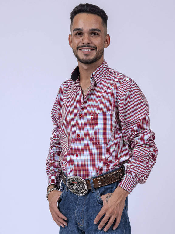 Um jovem está posando em pé, com um sorriso no rosto. Ele usa uma camisa de manga longa de cor rosa claro com pequenos padrões, calças jeans azuis e botas escuras. O jovem está usando um cinto com uma fivela grande e está em um fundo neutro, claro.