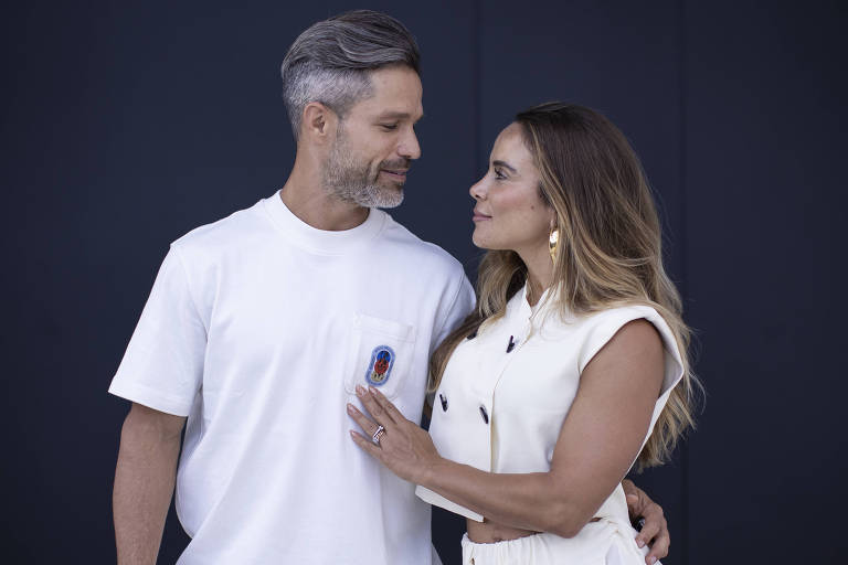 Um homem e uma mulher estão em uma pose romântica, olhando um para o outro. O homem usa uma camiseta branca e tem cabelo grisalho, enquanto a mulher veste um conjunto branco e tem cabelo longo e solto. Eles estão em um fundo escuro, que destaca suas roupas claras.